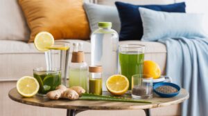 A round table with healthy morning drinks like green juice and lemon water sits beside ginger root, chia seeds, aloe leaf, halved lemons, and an orange. Glass bottles and a sofa with colorful cushions are in the background.