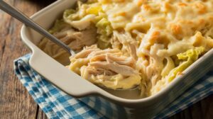 A casserole dish filled with cheesy chicken and noodle bake, perfect for meal prep, with a spoon lifting a portion. The dish sits on a blue checkered cloth on a wooden table.