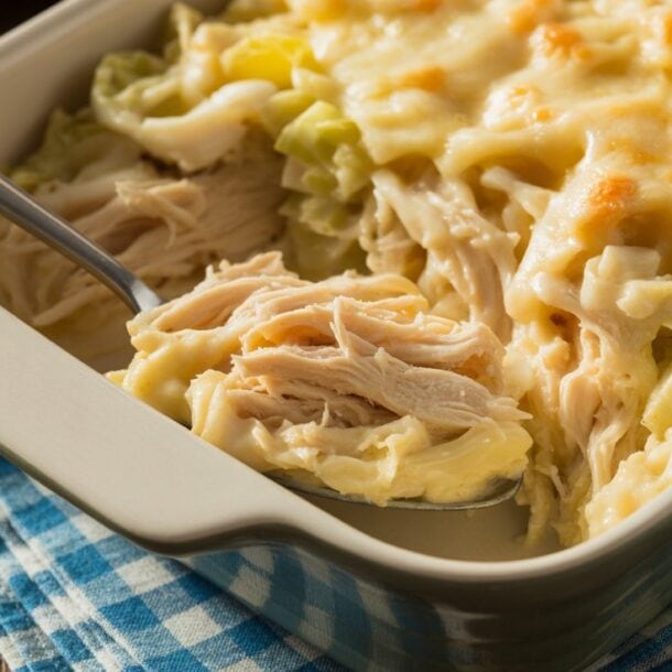 A casserole dish filled with cheesy chicken and noodle bake, perfect for meal prep, with a spoon lifting a portion. The dish sits on a blue checkered cloth on a wooden table.