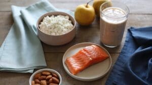 A wooden table displays a bowl of cottage cheese, a glass of smoothie, two yellow apples, a plate of raw salmon, and almonds—an ideal spread if you’re wondering how much protein should you eat daily. Light blue and navy napkins sit nearby.