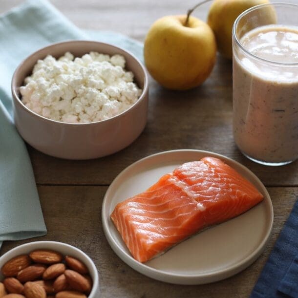 A wooden table displays a bowl of cottage cheese, a glass of smoothie, two yellow apples, a plate of raw salmon, and almonds—an ideal spread if you’re wondering how much protein should you eat daily. Light blue and navy napkins sit nearby.