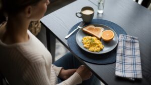 A person sits at a table with a plate of scrambled eggs, toast, and half an orange—natural ways to feel full—next to a mug of coffee and a glass of water. A fork and a blue-checked napkin are on the table.