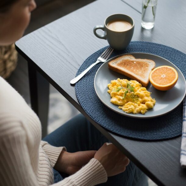 A person sits at a table with a plate of scrambled eggs, toast, and half an orange—natural ways to feel full—next to a mug of coffee and a glass of water. A fork and a blue-checked napkin are on the table.