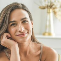A woman with long brown hair smiles gently, resting her chin on her hand. She is indoors with soft natural light and a blurred, elegant background featuring gold and white decor.