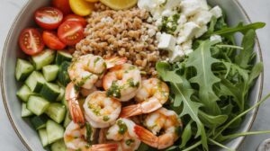 A colorful Farro Protein Bowl featuring lemon garlic shrimp with green sauce, cherry tomatoes, chopped cucumber, lemon slice, feta cheese, and fresh arugula arranged in sections for a high protein meal prep option.