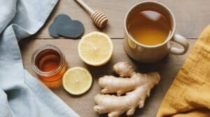 A mug of tea, a jar of honey with a honey dipper, fresh ginger root, and sliced lemon are arranged on a wooden table with blue and yellow cloth napkins—classic staples for natural cold remedies.