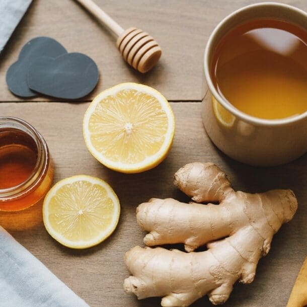 A mug of tea, a jar of honey with a honey dipper, fresh ginger root, and sliced lemon are arranged on a wooden table with blue and yellow cloth napkins—classic staples for natural cold remedies.