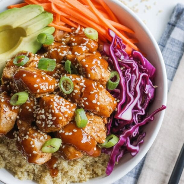 A Protein Bowl featuring quinoa, Spicy Honey Chicken glazed and topped with sesame seeds and green onions, sliced avocado, shredded carrots, and purple cabbage, served with a fork and napkin on the side.
