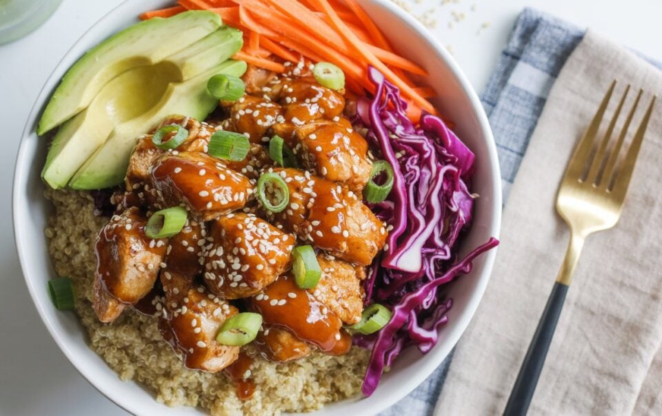 A Protein Bowl featuring quinoa, Spicy Honey Chicken glazed and topped with sesame seeds and green onions, sliced avocado, shredded carrots, and purple cabbage, served with a fork and napkin on the side.