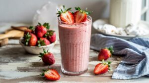 A glass of Strawberry Cheesecake Cottage Cheese Smoothie garnished with two halved strawberries, surrounded by fresh strawberries on a rustic table, with a bowl of strawberries and a blue striped cloth in the background.