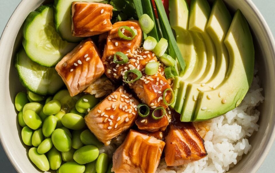 A vibrant rice bowl featuring teriyaki salmon cubes, avocado, cucumber, edamame, green onions, and sesame seeds—perfect for meal prep or a delicious lunch.