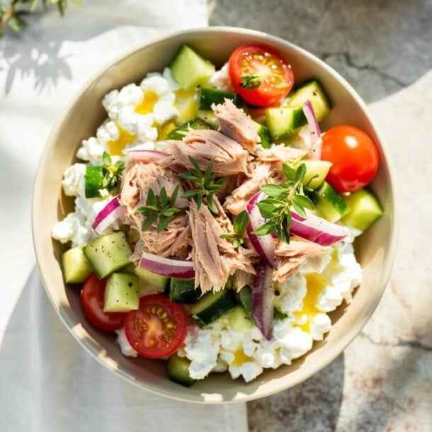 A Tuna Cottage Cheese Power Bowl filled with cucumber chunks, cherry tomato halves, red onion slices, and flaked tuna, garnished with fresh herbs, sits on a light surface with sunlight casting soft shadows.
