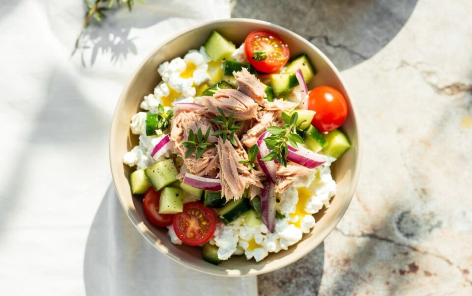 A Tuna Cottage Cheese Power Bowl filled with cucumber chunks, cherry tomato halves, red onion slices, and flaked tuna, garnished with fresh herbs, sits on a light surface with sunlight casting soft shadows.