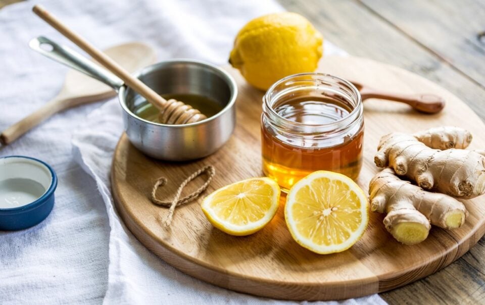 A wooden board with a jar of honey, sliced lemon, whole lemon, fresh ginger root, and a small bowl of honey with a dipper—classic ingredients for DIY Natural Cough Syrup—arranged on a white cloth on a rustic wooden table.