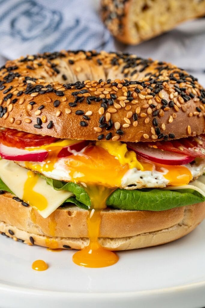 A greek yogurt bagel sandwich filled with leafy greens, cheese, tomato slices, and a fried egg with a runny yolk oozing onto the plate. Another greek yogurt bagel is blurred in the background.