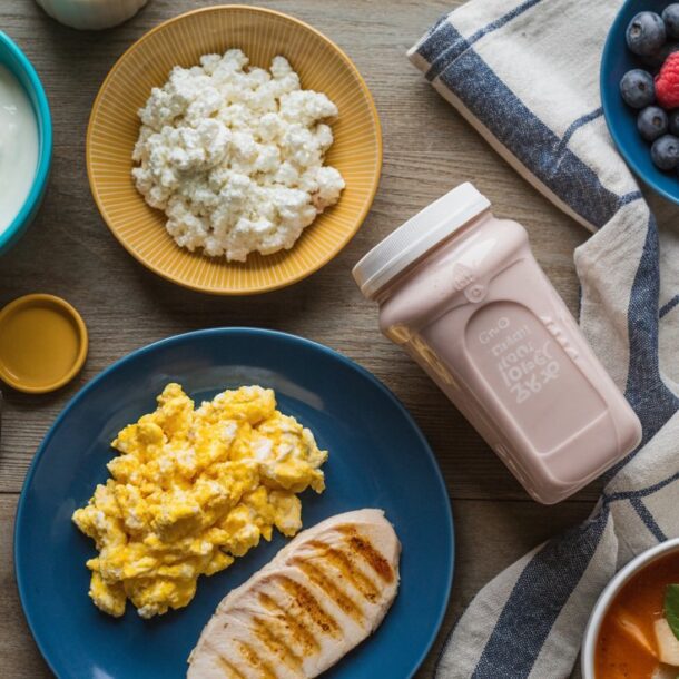A table set with healthy food swaps like scrambled eggs, grilled chicken, cottage cheese, yogurt, mixed berries, crackers, a pink Zepbound drink in a bottle, forks, and a striped napkin on a wooden surface.