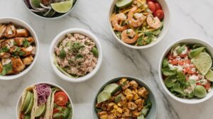 Seven bowls of assorted fresh High Protein Low Calorie Lunches on a marble surface, including shrimp salad, grilled chicken, tuna, tomatoes, avocado, lime wedges, and various greens, presented in a bright and appetizing arrangement.