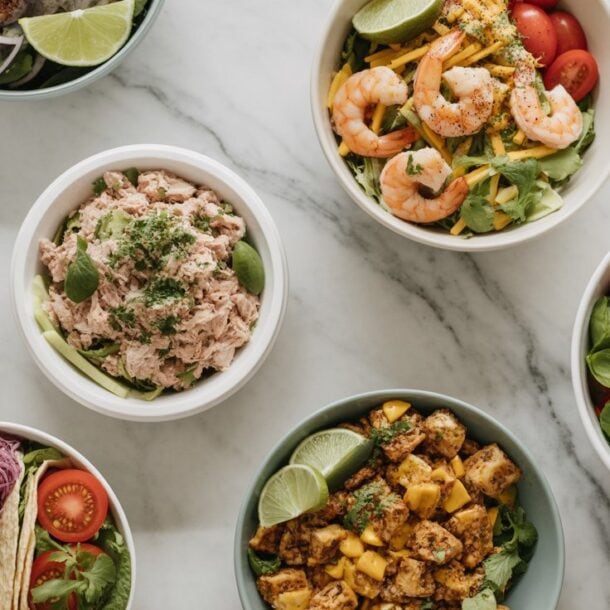 Seven bowls of assorted fresh High Protein Low Calorie Lunches on a marble surface, including shrimp salad, grilled chicken, tuna, tomatoes, avocado, lime wedges, and various greens, presented in a bright and appetizing arrangement.