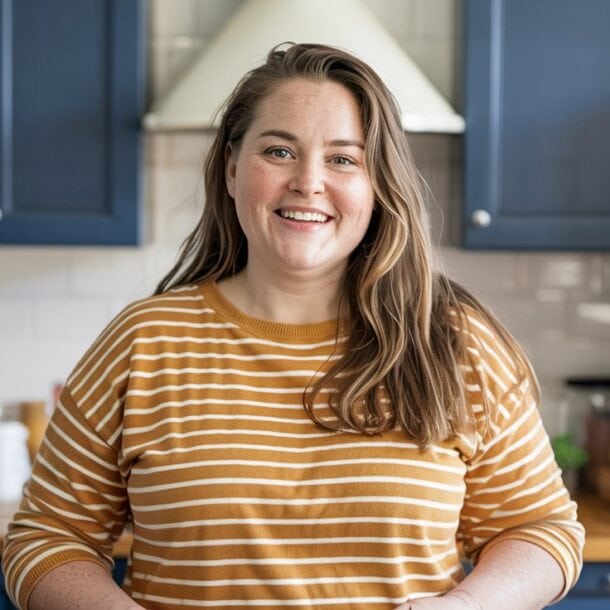 A woman with long brown hair, wearing a yellow and white striped shirt, smiles while standing in a kitchen with blue cabinets and a wooden countertop—ready to maximize results on her weight loss journey.