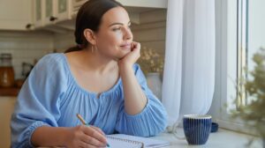 A woman in a light blue blouse sits at a kitchen table, resting her chin on her hand and looking thoughtfully out the window. Reflecting on her 110 pounds weight loss journey, she holds a pen over an open notebook, with a striped mug nearby.