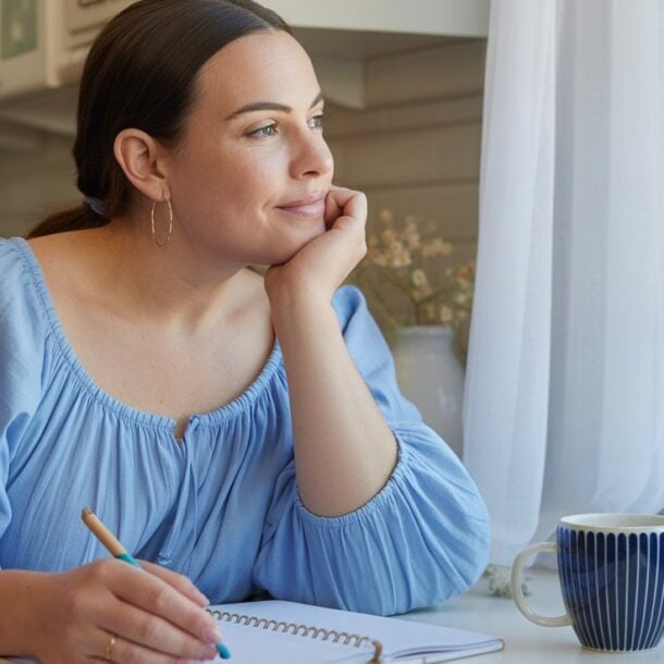 A woman in a light blue blouse sits at a kitchen table, resting her chin on her hand and looking thoughtfully out the window. Reflecting on her 110 pounds weight loss journey, she holds a pen over an open notebook, with a striped mug nearby.