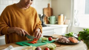 A person wearing a mustard yellow sweater slices vegetables on a green cutting board in a bright kitchen. Plates of rice, salmon—one of the best protein sources—and vegetables are on the counter nearby.