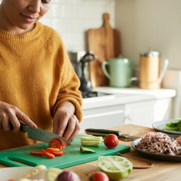 A person wearing a mustard yellow sweater slices vegetables on a green cutting board in a bright kitchen. Plates of rice, salmon—one of the best protein sources—and vegetables are on the counter nearby.