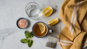 A cup of herbal tea with a lemon slice, a glass of water, lemon wedges, pink salt in a small bowl, fresh mint leaves, a tea bag, and a yellow cloth on a marble surface evoke the refreshing essence of a natural mounjaro drink.