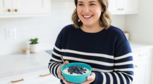 A smiling woman in a navy and white striped sweater holds a bowl of yogurt topped with fresh blueberries, enjoying a bright, modern kitchen as she follows the 30-30-30 Rule for a balanced morning routine.
