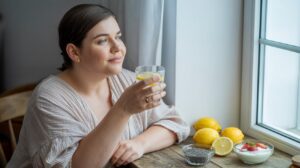 A woman sits at a wooden table by a window, holding a glass of lemon water and looking outside. On the table are lemons, a bowl of chia seeds—natural appetite suppressants—and a bowl of yogurt topped with berries.