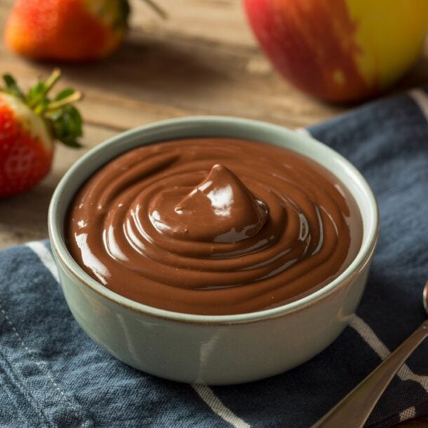 A bowl of creamy chocolate pudding sits on a blue napkin beside a spoon, with fresh strawberries and apples scattered on a rustic wooden table.