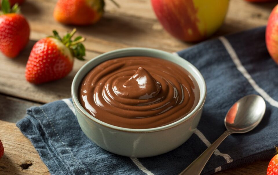A bowl of creamy chocolate pudding sits on a blue napkin beside a spoon, with fresh strawberries and apples scattered on a rustic wooden table.