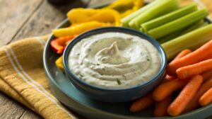 A plate of fresh vegetables, including celery sticks, baby carrots, and sliced peppers, surrounds a bowl of high-protein cottage cheese ranch dip garnished with herbs. The plate sits on a yellow-striped napkin on a wooden table.