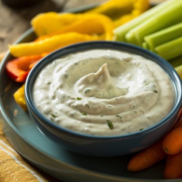 A plate of fresh vegetables, including celery sticks, baby carrots, and sliced peppers, surrounds a bowl of high-protein cottage cheese ranch dip garnished with herbs. The plate sits on a yellow-striped napkin on a wooden table.
