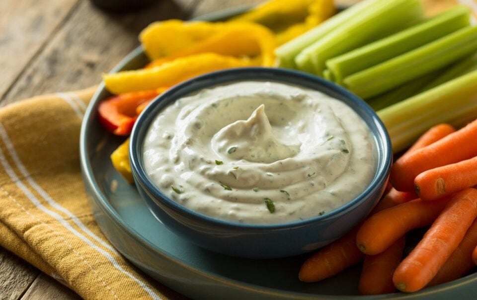A plate of fresh vegetables, including celery sticks, baby carrots, and sliced peppers, surrounds a bowl of high-protein cottage cheese ranch dip garnished with herbs. The plate sits on a yellow-striped napkin on a wooden table.