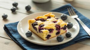 A slice of blueberry cake sits on a white plate with fresh blueberries scattered around, featuring a high protein cottage cheese blend, placed on a blue napkin with a fork nearby on a wooden table.