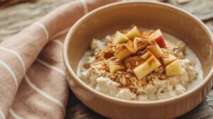 A bowl of oatmeal topped with diced apples, cinnamon, and oats sits on a rustic wooden table next to a striped cloth napkin, making it a perfect high fiber breakfast.