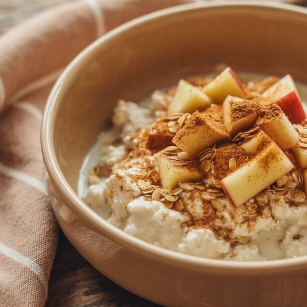 A bowl of oatmeal topped with diced apples, cinnamon, and oats sits on a rustic wooden table next to a striped cloth napkin, making it a perfect high fiber breakfast.