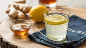 A glass of lemon water with lemon slices, a small bowl of honey, a whole lemon, and fresh ginger root on a wooden surface with a blue napkin—perfect for starting your 30-day lemon water challenge and enjoying the health benefits.