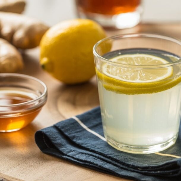 A glass of lemon water with lemon slices, a small bowl of honey, a whole lemon, and fresh ginger root on a wooden surface with a blue napkin—perfect for starting your 30-day lemon water challenge and enjoying the health benefits.