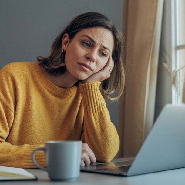 A woman in a yellow sweater sits at a table, looking thoughtfully at a laptop. A notebook, mug, and muffin—possibly chosen to curb afternoon food cravings—are on the table as sunlight comes through a nearby window.
