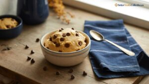 A bowl of Low Calorie cottage cheese cookie dough topped with chocolate chips sits on a wooden table beside a blue napkin, a spoon, and scattered chocolate chips. Another bowl is in the background.
