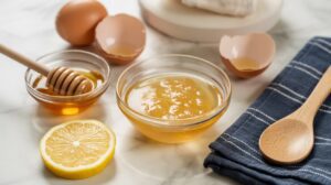 A bowl of honey, a wooden spoon, a lemon slice, eggshells, and a jar of honey with a dipper are arranged on marble—ingredients perfect for creating a natural Face Tightening Mask—next to a dark striped cloth.