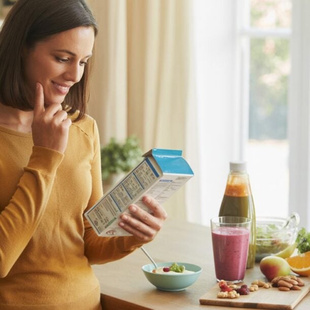 A woman in a mustard sweater stands in a kitchen, smiling as she reads a cereal box. On the counter are a bowl of cereal, dessert, nuts, fruit, and juice—perfect for supporting her body and balanced blood sugar.