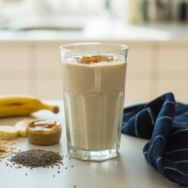A glass of creamy breakfast smoothie sits on the counter, surrounded by a banana, oats, chia seeds, and a spoonful of nut butter—perfect for a protein fiber boost. A blue striped cloth adds charm to the bright kitchen setting.