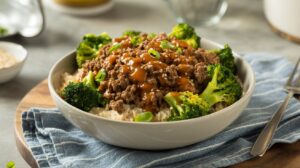 A Beef & Broccoli Bowl features white rice topped with ground beef in a savory brown sauce, garnished with green onions and surrounded by steamed broccoli, all resting on a blue striped cloth.