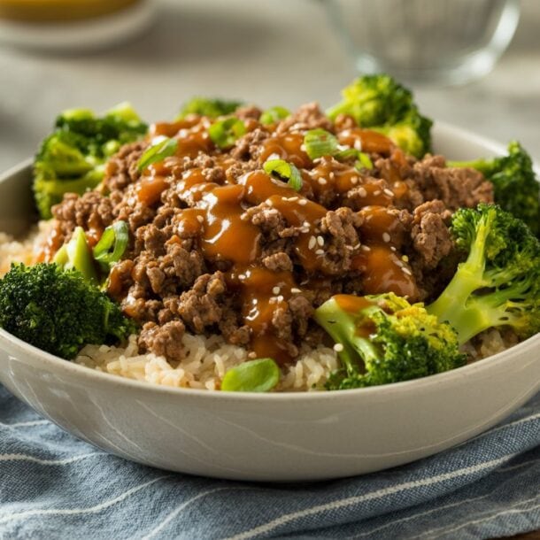 A Beef & Broccoli Bowl features white rice topped with ground beef in a savory brown sauce, garnished with green onions and surrounded by steamed broccoli, all resting on a blue striped cloth.