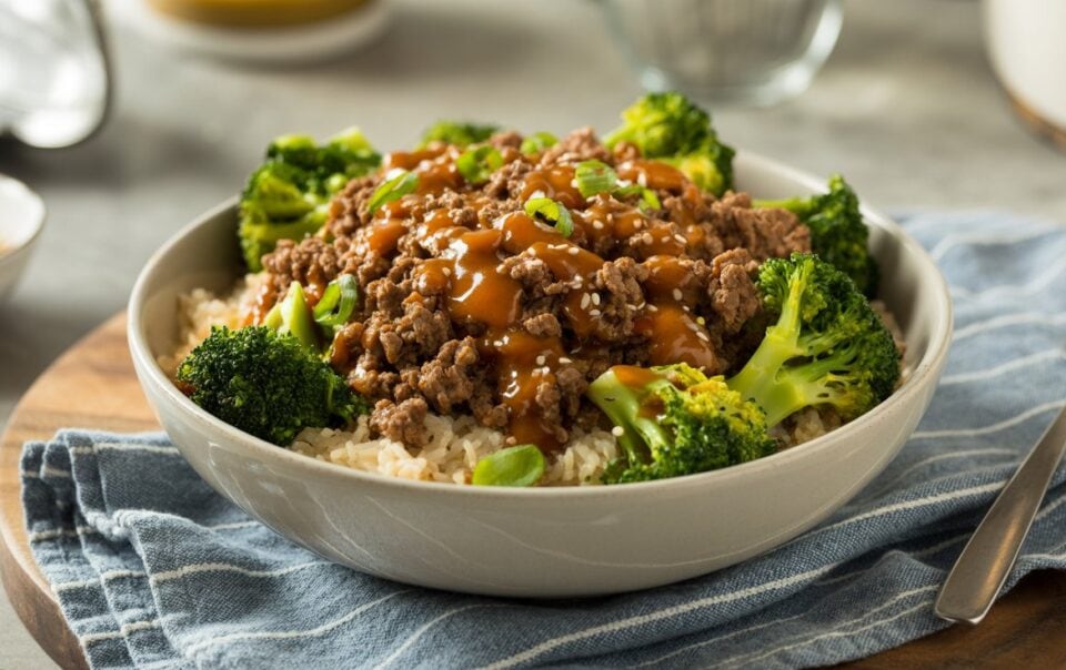A Beef & Broccoli Bowl features white rice topped with ground beef in a savory brown sauce, garnished with green onions and surrounded by steamed broccoli, all resting on a blue striped cloth.