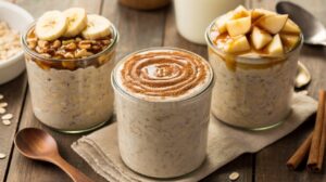 Three jars of overnight oats recipe are on a wooden table: one topped with banana slices and nuts, one with a swirl of cinnamon, and one with chopped apples, caramel sauce, and brown sugar. A wooden spoon and cinnamon sticks are nearby.