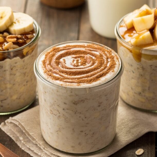 Three jars of overnight oats recipe are on a wooden table: one topped with banana slices and nuts, one with a swirl of cinnamon, and one with chopped apples, caramel sauce, and brown sugar. A wooden spoon and cinnamon sticks are nearby.